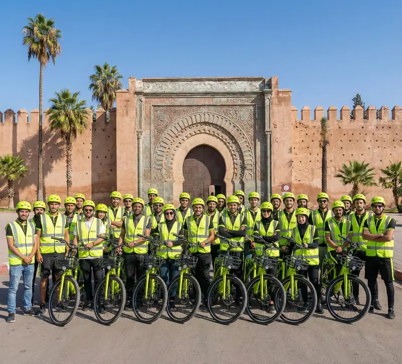 A large group of riders in lime safety vests posing with their bikes in front of a Marrakech city gate